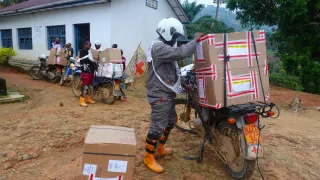 DRC-Health-1 Des travailleurs humanitaires chargent de grandes boîtes de matériel médical sur des motos devant un centre de santé rural, en préparation pour la livraison d’aide dans des zones reculées