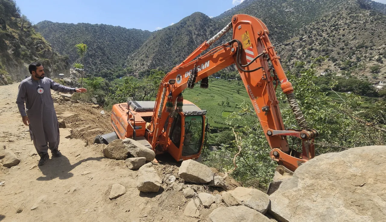 Afghanistan earthquake August 25 A man wearing a Red Crescent emblem points towards an excavator off a dirt road on a steep hillside in a mountainous area of Afghanistan, following the earthquake.