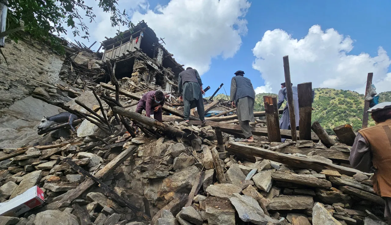 Afgh_1_20250904_133200 People search through the rubble of collapsed buildings after an earthquake, surrounded by broken wooden beams and stones, with mountains visible in the background under a partly cloudy sky.