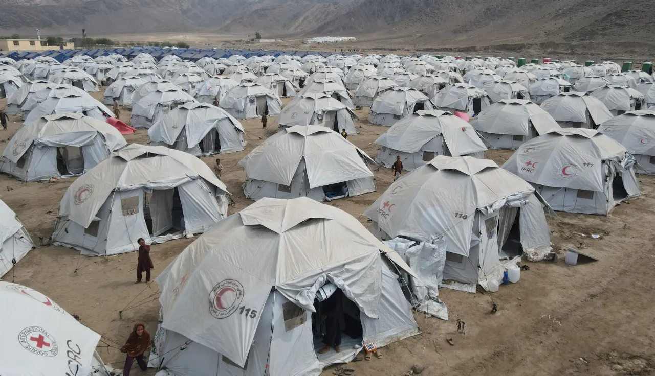 Afgh_2_Camp Rows of white humanitarian relief tents with Red Cross and Red Crescent logos set up in a camp on dusty ground, surrounded by mountains in the background.