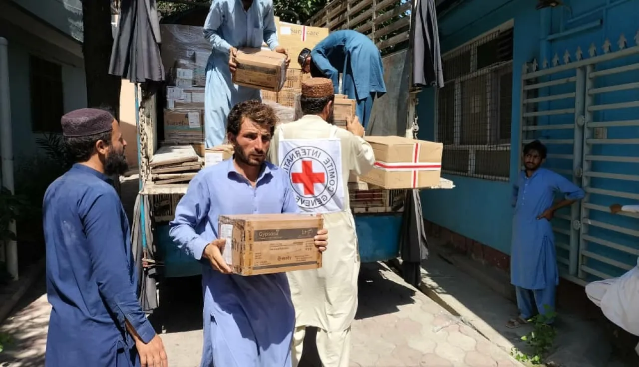 Afgh_3_Kunar-HSP Humanitarian workers unload boxes of supplies from a truck, with one man wearing a vest marked with the Red Cross emblem, distributing aid outside a building.