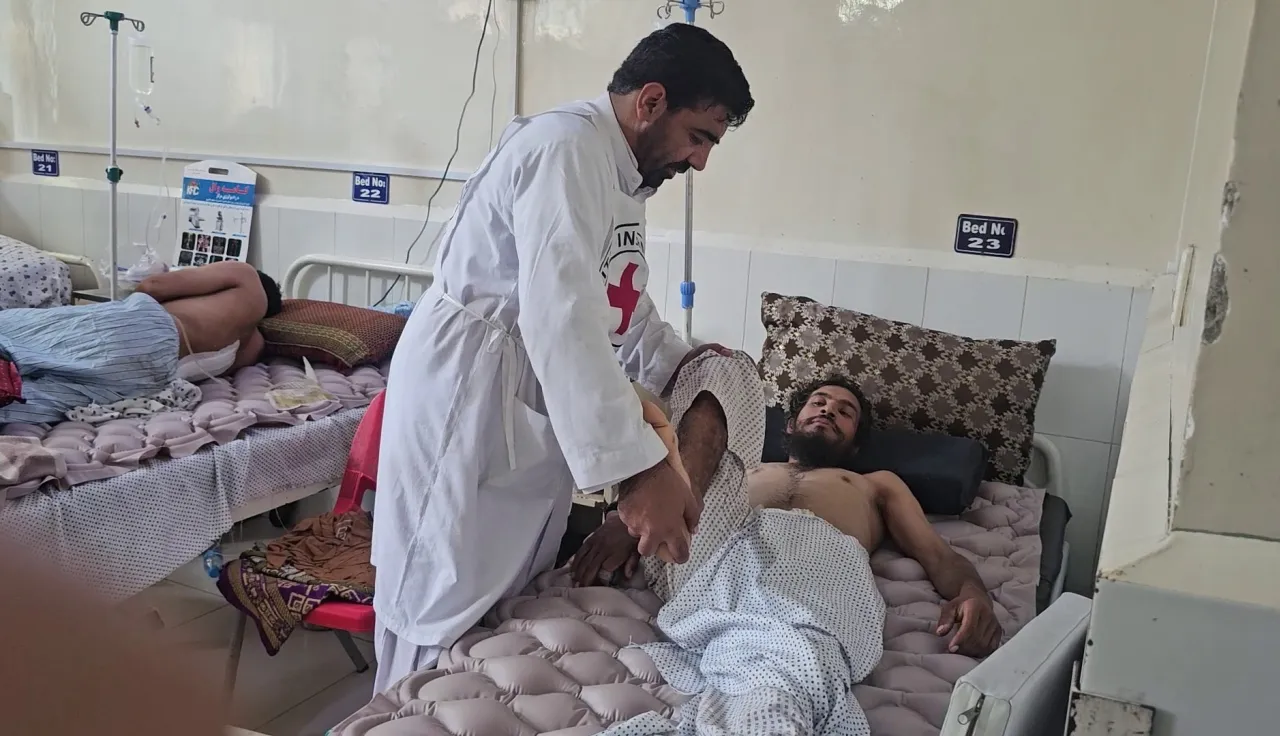 Afgh_4_JAL-PRC A medical worker in a white coat assists an injured man lying on a hospital bed, with other patients resting on beds in the background.