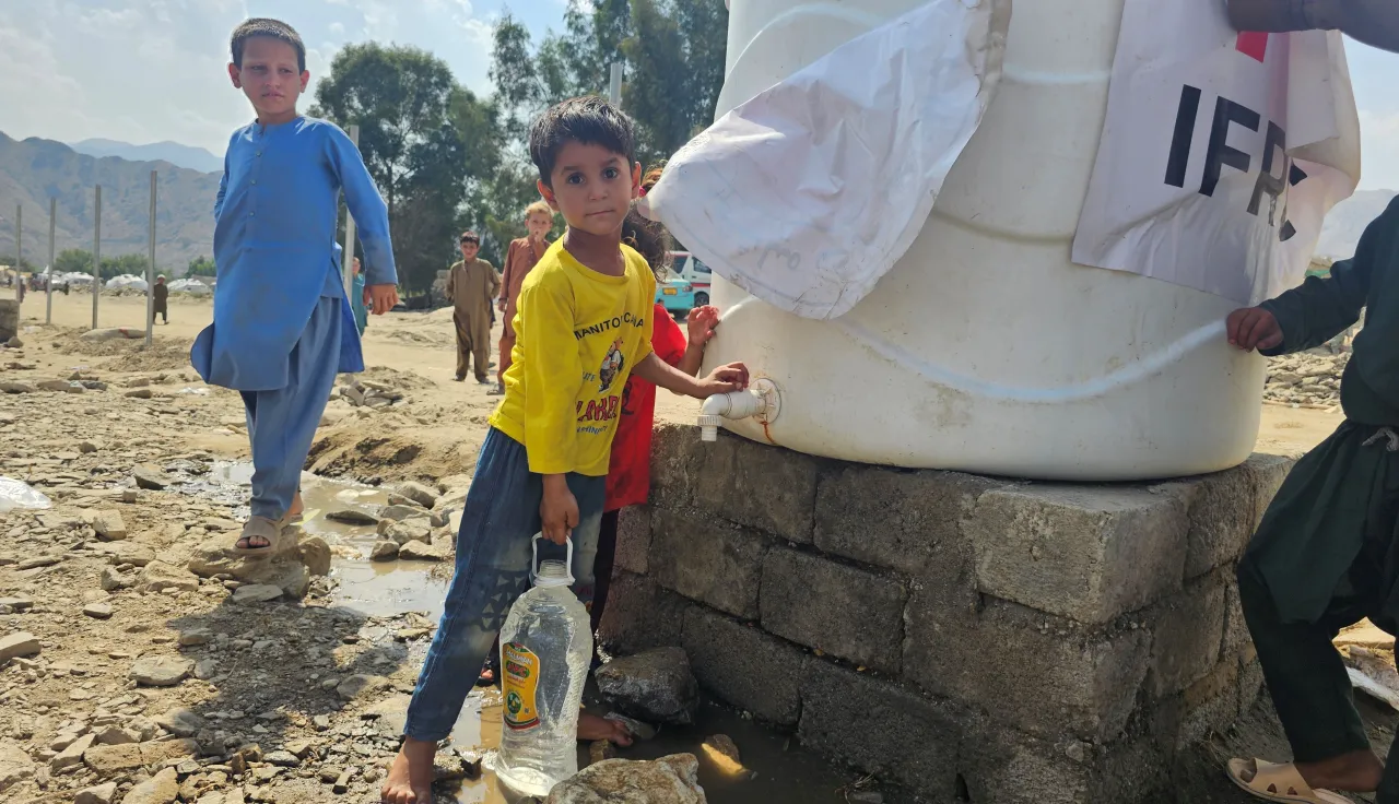 Afgh_7_P-1--18---1- Children collect clean water from a large tank marked with humanitarian logos, one boy filling a plastic container, in a dry outdoor setting with mountains in the background.