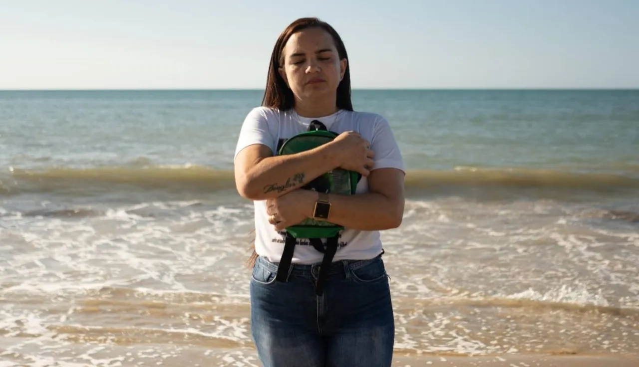 Una mujer aferrada a una mochila con los ojos cerrados, de espaldas al mar.