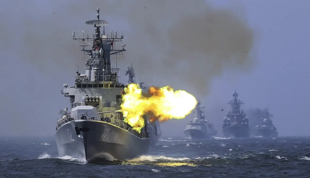 A ship at sea firing artillery while three other ships follow behind.
