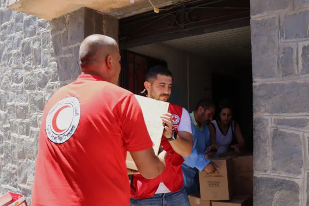 Syria Sweida delivering supplies Humanitarian workers wearing Red Crescent and Red Cross vests unload and distribute boxes of aid supplies at a building entrance.