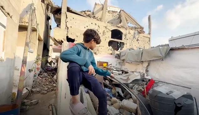Gaza-AN-news-boy-home A young boy sits on a concrete ledge amid the rubble of a destroyed building, looking out over the debris and damage around him under a partly cloudy sky.