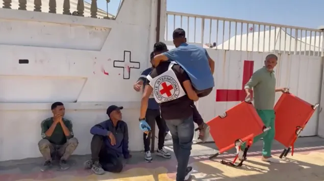 Gaza-AV-news-field-hospital A man carries another person on his back toward a gate marked with a red cross, while others sit or stand nearby, including a man holding two orange stretchers.