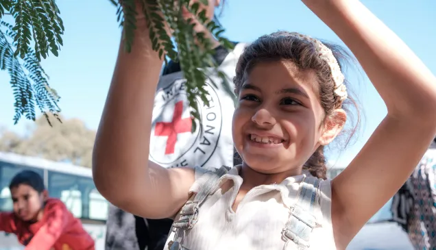 Batoul plays with a tree branch, smiling, with blue sky and a red cross in the background