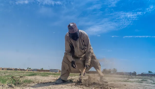 Nigeria Maidguri - DSC01663 Churi Ibrahim, a 70-year-old farmer, works the dry, dusty soil with a hoe under a bright blue sky in a rural area, with modest shelters visible in the background — a scene reflecting resilience in the face of harsh living conditions.