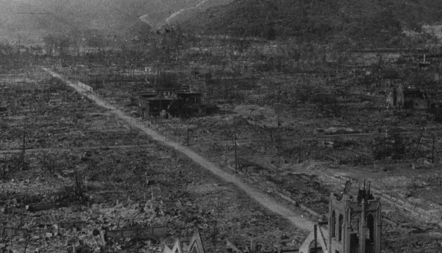 Hiroshima Nakaku. After the explosion of the atom bomb. A black and white photograph showing the devastated landscape of Hiroshima after the nuclear bombing in August 1945. The area is flattened and burned, with only a few skeletal buildings and bare trees remaining. A straight dirt road runs through the center, surrounded by rubble and destruction, with mountains visible in the background.