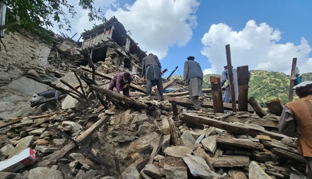 Afgh_1_20250904_133200 People search through the rubble of collapsed buildings after an earthquake, surrounded by broken wooden beams and stones, with mountains visible in the background under a partly cloudy sky.