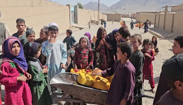 A group of children gather around a wheelbarrow carrying yellow jerrycans in a dusty street, with buildings and mountains in the background.