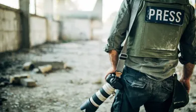 Pessoa com colete escrito "press" segura uma máquina fotográfica num edifício destruído.