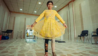 طفلة في فستان أصفر تقف على قدميها وإحدى ساقيها صناعية تبتسم وتمسك طرفي الفستان بيديها
