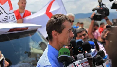 An ICRC press conference, with a man at a bank of microphones, a huddle of press and a camera, and ICRC staff and flags behind him