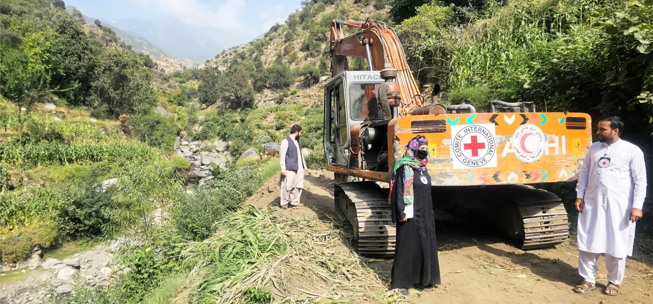 Afgh_5_Heavy-Machinery People stand near an excavator marked with Red Cross logos, working on clearing or repairing a rural road in a green, mountainous area.