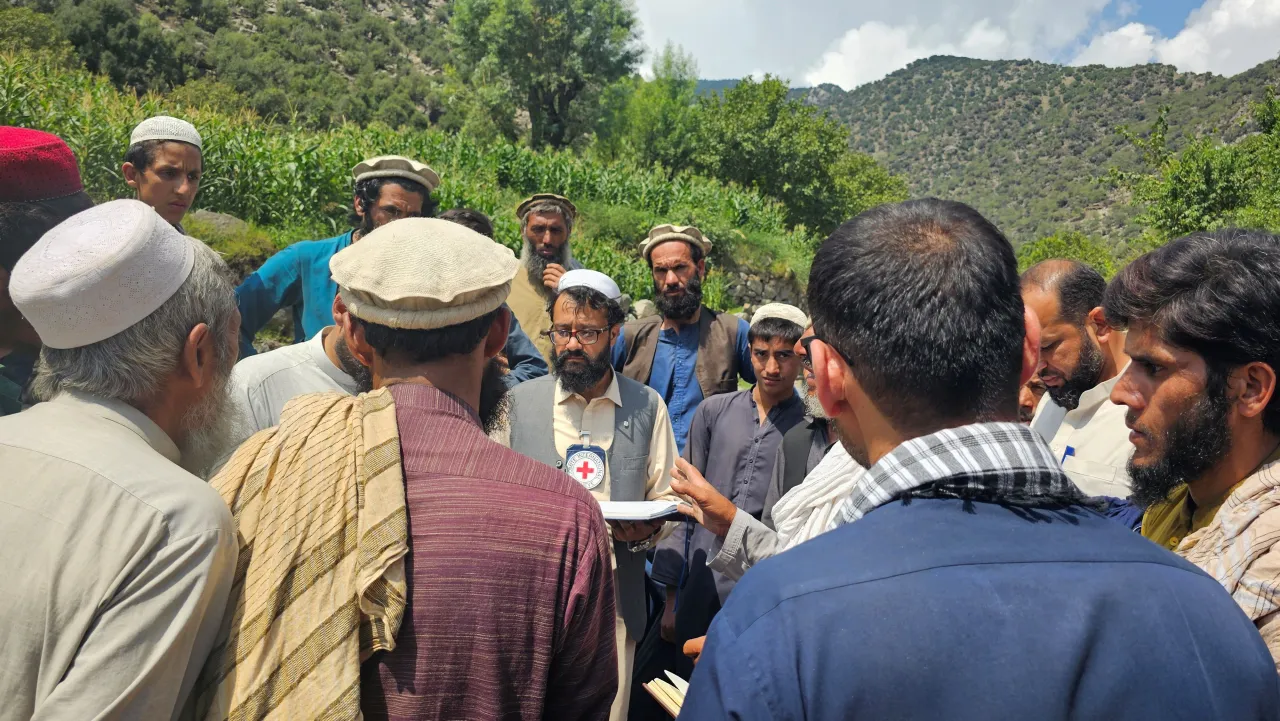 Afgh_6_20250908_105158 A group of men gather outdoors in a rural, mountainous area, listening to a humanitarian worker wearing a vest with a Red Cross emblem.
