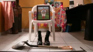 Una foto de una mujer sosteniendo un retrato está apoyada sobre una silla. A los pies de la silla, unas botas y elementos de trabajo como una pala y un machete.