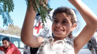 Lebanon IDP Batoul 1 Batoul plays with a tree branch, smiling, with blue sky and a red cross in the background