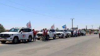 Syria Sweida convoy A convoy of white vehicles marked with Red Cross emblems and flags parked on a road under a clear blue sky, with humanitarian workers standing beside them.