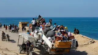 A crowded truck loaded with people and belongings drives along a coastal road, as others walk or travel on carts beside the sea.
