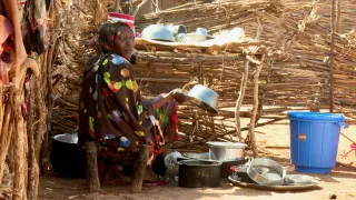 A woman washes dishes in an outdoor area surrounded by wooden fencing, with metal pots and a blue plastic bucket nearby.