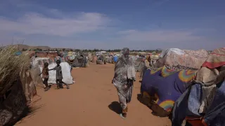 A woman walks through a dusty displacement site in Tawila, Sudan, passing makeshift shelters covered with tarps and fabric, while groups of people stand and move between the crowded structures under a clear blue sky.