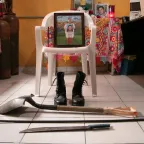 Una foto de una mujer sosteniendo un retrato está apoyada sobre una silla. A los pies de la silla, unas botas y elementos de trabajo como una pala y un machete.