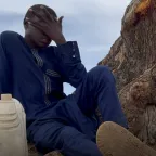 Nigeria food security A young boy in a navy blue outfit sits at the base of a large tree, covering his face with one hand in a gesture of distress or exhaustion. A plastic water container rests beside him.