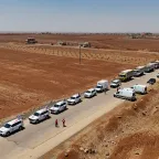 Sweida Syria July 2025 A long humanitarian aid convoy stretches along a rural road in Sweida, Syria, in July 2025. The convoy includes dozens of white vehicles marked with the Red Cross and Red Crescent emblems, along with large trucks carrying relief supplies. The road runs through a dry, reddish-brown landscape with sparse vegetation and scattered buildings in the distance. Aid workers in red uniforms walk near the vehicles under a clear blue sky.