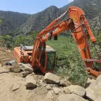 Afghanistan earthquake August 25 A man wearing a Red Crescent emblem points towards an excavator off a dirt road on a steep hillside in a mountainous area of Afghanistan, following the earthquake.