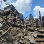 Afgh_1_20250904_133200 People search through the rubble of collapsed buildings after an earthquake, surrounded by broken wooden beams and stones, with mountains visible in the background under a partly cloudy sky.