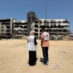 Gaza-July-2025 Two humanitarian workers wearing vests with Red Cross and partner organization logos stand in front of a heavily damaged, partially destroyed building, assessing the destruction in Gaza
