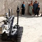 A robot used in war at the foreground while children look on in the background. 