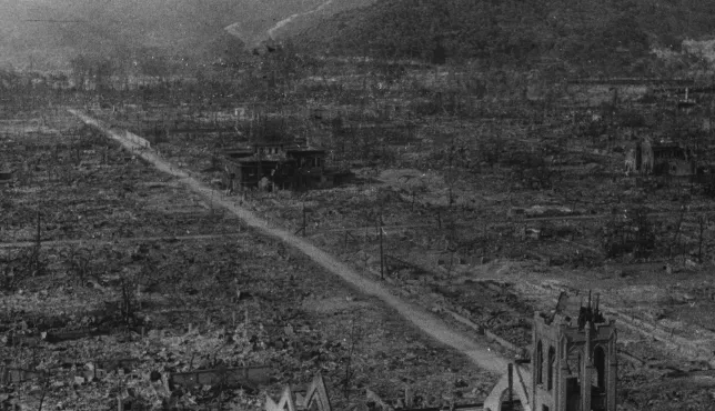 Hiroshima Nakaku. After the explosion of the atom bomb. A black and white photograph showing the devastated landscape of Hiroshima after the nuclear bombing in August 1945. The area is flattened and burned, with only a few skeletal buildings and bare trees remaining. A straight dirt road runs through the center, surrounded by rubble and destruction, with mountains visible in the background.